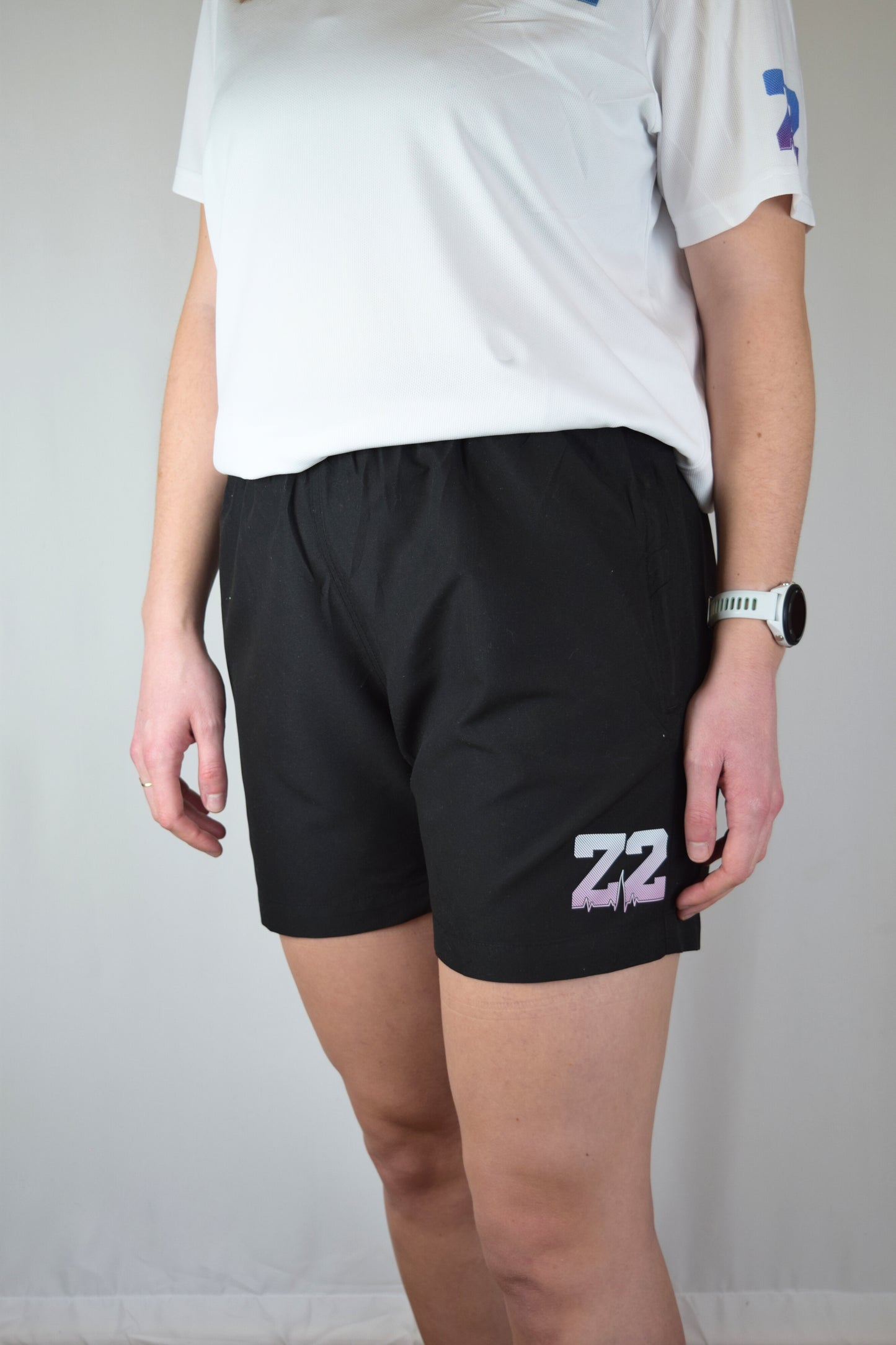 Lady wearing comfortable black athletic shorts with 'Z2' logo and a white shirt with a visible brand logo on a plain background